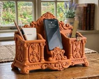 May include: A terracotta-colored desk organizer featuring ornate relief carvings, a central smartphone stand, and two side compartments. The organizer holds a small notepad, a pen, a pencil, and a ruler, displaying a detailed floral pattern.