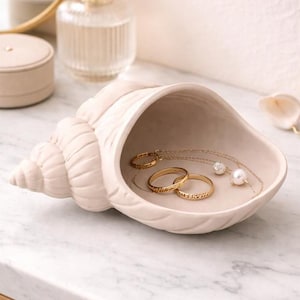 May include: A cream-colored seashell-shaped jewelry dish holding gold rings and a necklace with a pearl pendant. The dish sits on a marble surface, with a perfume bottle and small round container in the background.