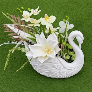 Puede incluir: Una maceta con forma de cisne blanco llena de un arreglo de flores artificiales, que incluye una gran magnolia blanca, orquídeas blancas más pequeñas, follaje verde y tallos decorativos plumosos blancos y marrones en el cuerpo.
