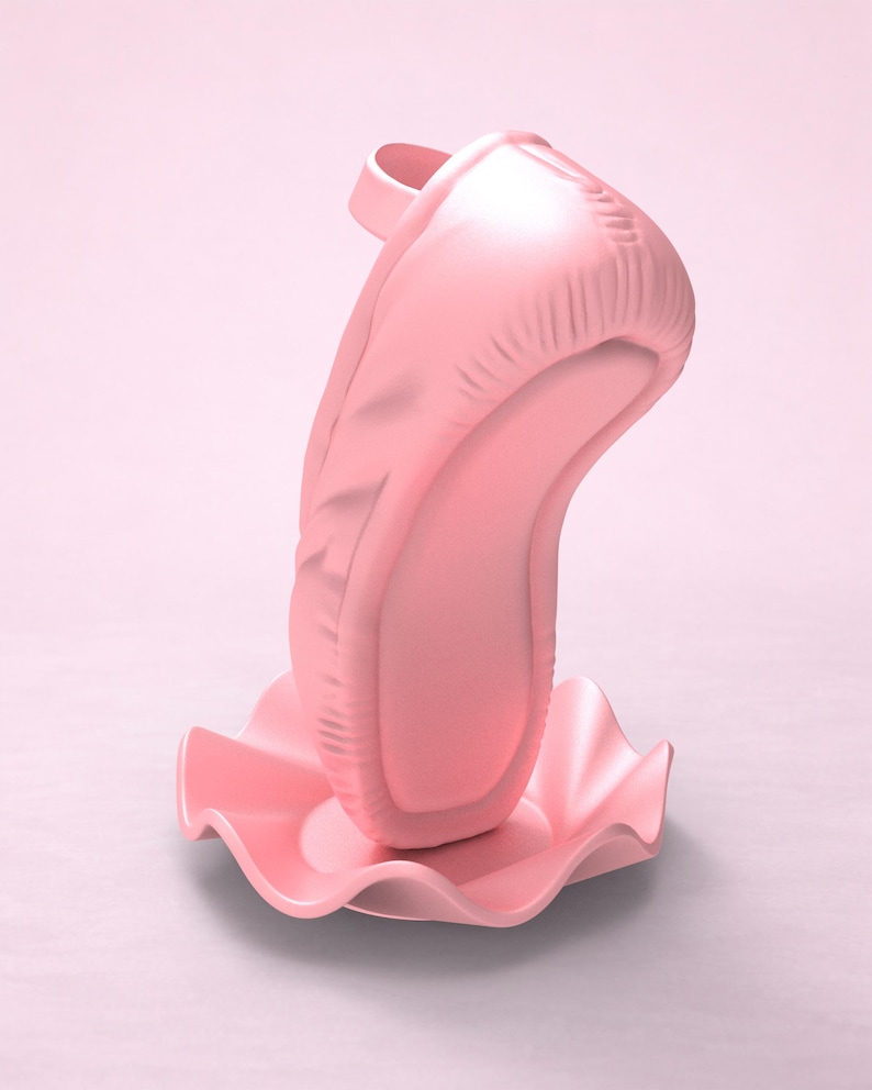 May include: A light pink ballet slipper standing vertically on a matching pink circular base with a ruffled, wavy edge. The shoe features a smooth, matte finish with visible stitching details along the heel and a small strap at the top opening.