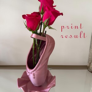 May include: A pink ballet slipper shaped vase with a small bow detail at the toe, holding three vibrant dark pink roses. The text print result is visible in red script to the right of the vase, which sits on a matching pink ruffled base.