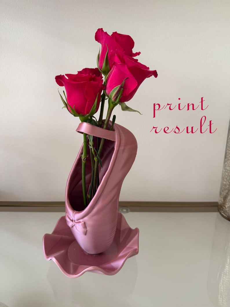 May include: A pink ballet slipper shaped vase with a small bow detail at the toe, holding three vibrant dark pink roses. The text print result is visible in red script to the right of the vase, which sits on a matching pink ruffled base.