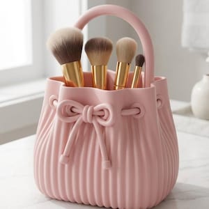 May include: A pink, ribbed cosmetic brush holder with a handle, holding several makeup brushes. The brushes have gold-colored metal handles and soft bristles. The holder has a bow detail on the front.