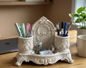 May include: A cream-colored desk organizer featuring two cylindrical pen holders flanking a central back panel with ornate shell and acanthus leaf carvings. The base has decorative scrollwork feet and a central tray holding a small glass jar.