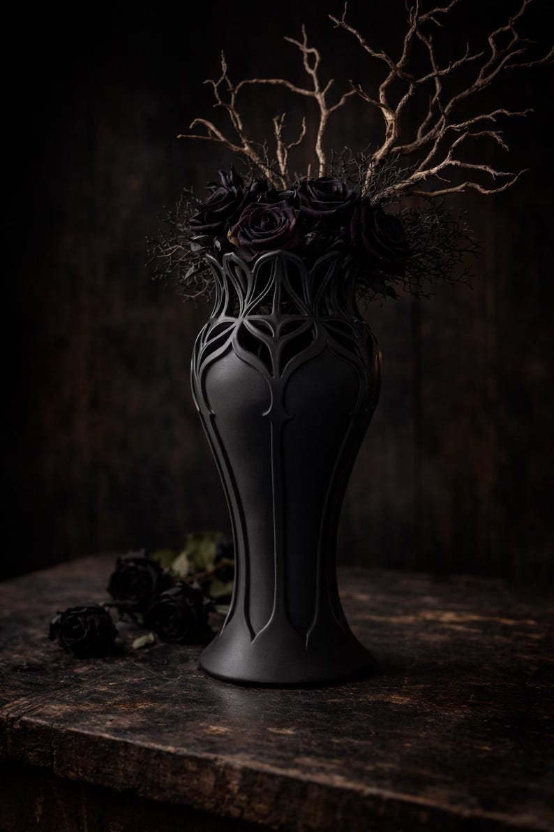 May include: A matte black vase with an intricate, openwork design, holding a bouquet of dark roses and golden branches. The vase sits on a dark wooden surface, with a few loose roses nearby. The overall aesthetic is gothic and elegant.