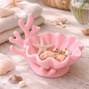 Pode incluir: Uma concha rosa de cerâmica com detalhes em forma de coral, contendo uma estrela do mar, conchas e joias. Sobre uma superfície de madeira clara, com mais estrelas do mar e conchas.