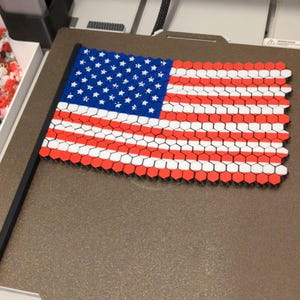 Op de afbeelding: Een 3D-geprinte Amerikaanse vlag met een zwarte paal. De vlag is gemaakt van rode, witte en blauwe zeshoekige tegels. Het blauwe veld heeft witte sterren en de strepen wisselen af tussen rood en wit.