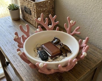 May include: A white ceramic bowl featuring a decorative pink coral-like rim adorned with small white spherical beads. The bowl contains a brown leather wallet, a pair of dark-framed sunglasses, a set of metal keys, and several loose coins.