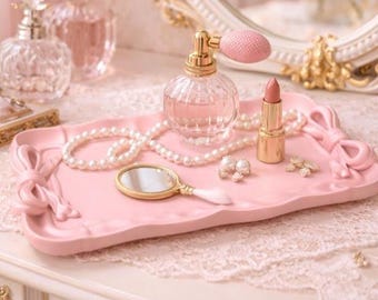 May include: A pink vanity tray with bow details holds a pearl necklace, a small hand mirror, earrings, and a lipstick. A perfume bottle with a pink atomizer and other perfume bottles are in the background. The scene is set on a lace-covered surface.
