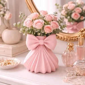 Peut inclure: Un vase rose en céramique en forme de nœud, rempli de roses roses et de verdure. Le vase est posé sur une surface blanche, avec un miroir et d'autres objets décoratifs en arrière-plan. Thème romantique et féminin.