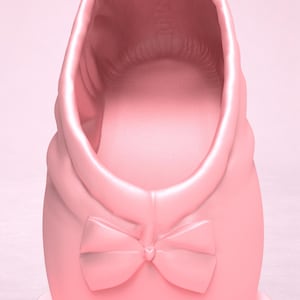 May include: A light pink ballet slipper featuring a smooth, matte finish and a decorative bow centered at the front. The shoe has a rounded opening and a structured design, showcasing a uniform pastel pink color throughout the entire exterior surface.