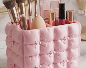 May include: A light pink rectangular makeup organizer with a quilted texture and small bow accents. The container holds several makeup brushes with rose gold handles, a beige makeup sponge, an eyeshadow palette, and three lipstick tubes.