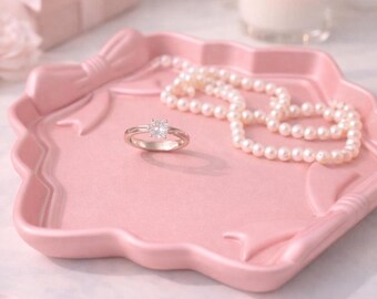 May include: A pink ceramic tray with a bow design holds a silver ring with a square gemstone and a strand of pearls. A lit candle and a pink gift box are in the background, creating a soft, romantic aesthetic.