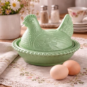 Könnte beinhalten: Ein hellgrüner Eierhalter in Form einer Henne aus Keramik mit strukturierter Oberfläche, der auf einem passenden Sockel steht. Zwei braune Eier liegen neben dem Halter auf einer Tischdecke mit Blumenmuster. Eine Vase mit weißen Blumen befindet sich im Hintergrund.