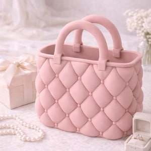 May include: A pink handbag with a quilted design and two rounded handles. The bag is sitting on a white surface, next to a gift box, a pearl necklace, and a ring box.