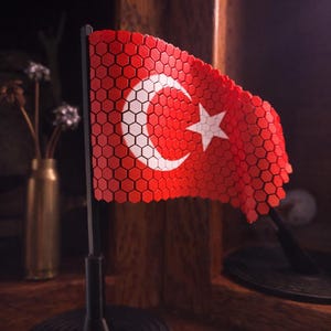 Può includere: Una bandiera turca rossa e bianca, con una luna crescente e una stella, è esposta su un supporto nero. La bandiera presenta un esclusivo design a nido d'ape. Lo sfondo è in legno scuro.