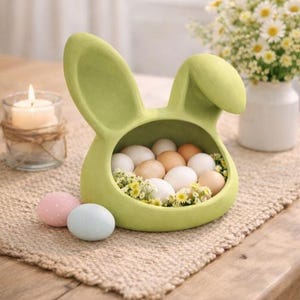 May include: A lime green bunny-shaped container filled with eggs and small yellow flowers. Two pastel-colored eggs sit in front of the container. A lit candle and a vase of daisies are in the background. The container is on a woven mat.