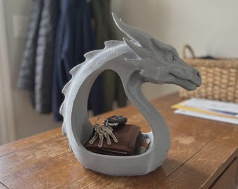 May include: A light gray, 3D-printed dragon-shaped bowl with a curved neck and head. The hollow center of the dragon serves as a storage compartment, currently holding a brown leather wallet, a set of metal keys, and a black car key fob.