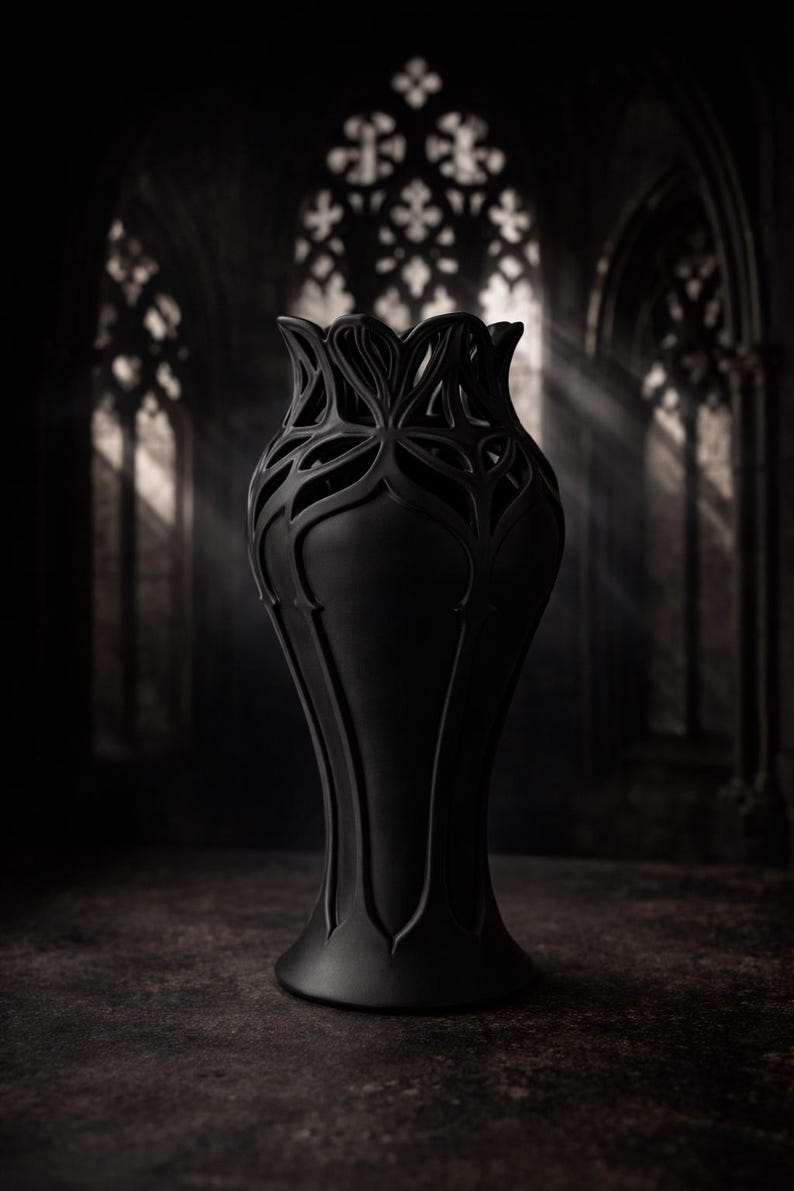May include: A tall, black vase with a unique, openwork floral design at the top. The vase has a slender, elongated shape and sits on a flared base. The background is a dark, gothic-style interior with arched windows.