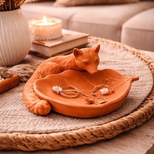 Puede incluir: Una taza de cerámica blanca con asa negra y el texto "But First, Coffee" en negrita negra. La taza se encuentra sobre un fondo borroso, sugiriendo un ambiente acogedor.