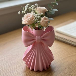 Puede incluir: Un jarrón de cerámica rosa con forma de lazo, que contiene rosas rosas claras y pequeñas flores blancas. El jarrón tiene un diseño plisado en la parte inferior. El arreglo está sobre una superficie de madera, cerca de una ventana y un cuaderno.