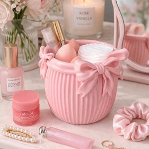 Può includere: Una scena di vanità rosa con prodotti di bellezza. Un contenitore rosa a coste con un fiocco contiene spugnette per il trucco e dischetti di cotone. Altri articoli includono un balsamo per le labbra, un elastico per capelli, una forcina e una candela "Rose Vanilla".