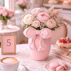 Puede incluir: Un jarrón rosa con forma de lazo contiene un ramo de rosas rosas y crema y paniculata. Una pequeña tarta con fresas, macarons y una taza de café están sobre la mesa. Un número 5 enmarcado también está sobre la mesa.