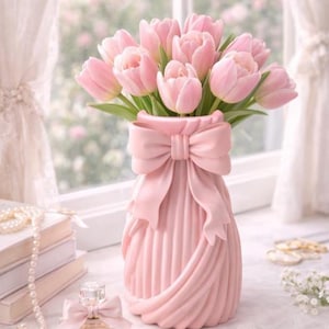 Puede incluir: Un ramo de tulipanes rosas en un jarrón rosa a juego con un diseño de lazo. El jarrón tiene una textura acanalada. También se ve un frasco de perfume y libros, cerca de una ventana.