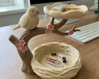 May include: A desk organizer featuring a light-colored bird perched on a brown branch with pink flowers. The branch supports a small, round tray at the top and a woven-style nest base at the bottom, both designed to hold small office supplies.