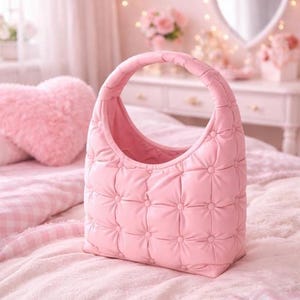 May include: A pink quilted handbag with a rounded handle. The bag has a grid pattern of stitched squares and is set on a pink bedspread. The background includes a heart-shaped mirror and a fluffy heart-shaped pillow.