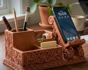 May include: A terracotta desk organizer featuring intricate floral relief carvings, multiple compartments for pens and office supplies, and an integrated smartphone stand displaying a screen with the time 10:09 and various app icons.