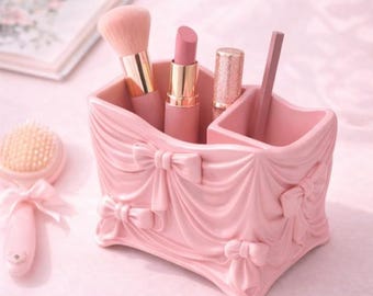 May include: A pink makeup organizer with a draped fabric design and bow accents. The organizer holds a makeup brush, lipstick, and other beauty products. A small heart-shaped item and a brush are also visible.