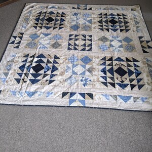 May include: A large, handmade quilt with a geometric pattern in shades of blue, tan, and black. The quilt features squares and triangles arranged in a repeating design. The quilt is laid out on a gray carpet.