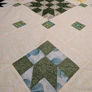 May include: Close-up of a handmade quilt with a diamond pattern. The quilt features various green, yellow, and white floral and geometric designs. The quilt is made of fabric with a soft, quilted texture.