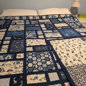 Peut inclure: Une courtepointe patchwork bleue et blanche, orn&eacute;e de motifs vari&eacute;s tels que des poissons, des fleurs et des formes g&eacute;om&eacute;triques. La courtepointe est pos&eacute;e sur un lit, mettant en valeur ses blocs rectangulaires et ses bordures bleu fonc&eacute;. L'ensemble est un m&eacute;lange de styles traditionnels et modernes.