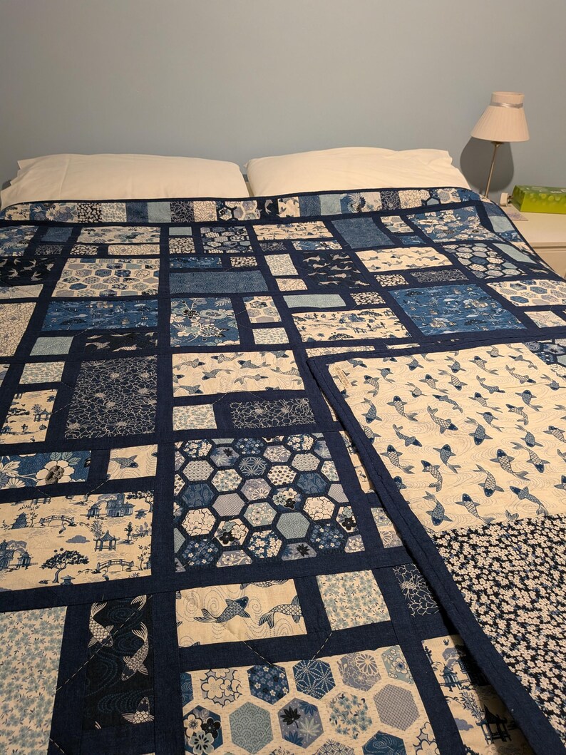 Peut inclure: Une courtepointe patchwork bleue et blanche, orn&eacute;e de motifs vari&eacute;s tels que des poissons, des fleurs et des formes g&eacute;om&eacute;triques. La courtepointe est pos&eacute;e sur un lit, mettant en valeur ses blocs rectangulaires et ses bordures bleu fonc&eacute;. L'ensemble est un m&eacute;lange de styles traditionnels et modernes.