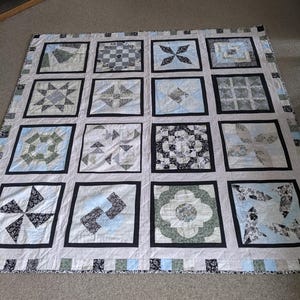 Op de afbeelding: Een handgemaakte quilt met een rasterpatroon van zestien vierkanten. Elk vierkant heeft een uniek geometrisch ontwerp in tinten blauw, grijs en wit, omlijst door zwarte randen. De rand van de quilt heeft een patroonrand.