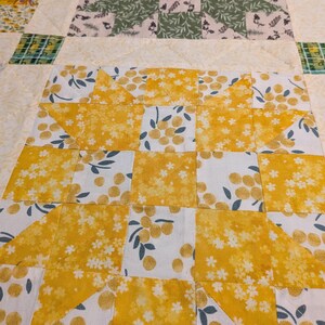 May include: Close-up of a handmade quilt with a patchwork design. The quilt features squares of yellow floral fabric, white fabric with yellow berries, and green and white patterned fabric. The quilt is a mix of colours and patterns.