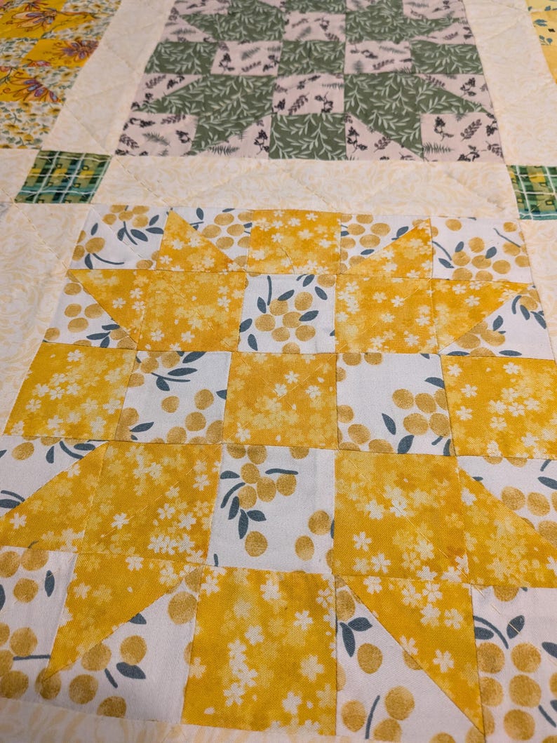 May include: Close-up of a handmade quilt with a patchwork design. The quilt features squares of yellow floral fabric, white fabric with yellow berries, and green and white patterned fabric. The quilt is a mix of colours and patterns.