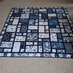 Peut inclure: Une courtepointe faite &agrave; la main avec un motif patchwork dans les tons de bleu et de blanc. La courtepointe pr&eacute;sente divers motifs g&eacute;om&eacute;triques, notamment des carr&eacute;s, des rectangles et des formes hexagonales. La courtepointe est bord&eacute;e d'un tissu bleu fonc&eacute;.