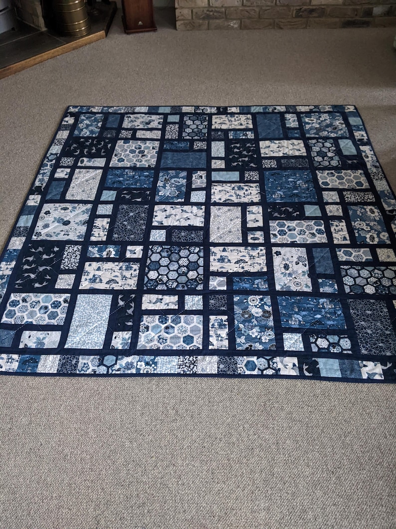 Peut inclure: Une courtepointe faite &agrave; la main avec un motif patchwork dans les tons de bleu et de blanc. La courtepointe pr&eacute;sente divers motifs g&eacute;om&eacute;triques, notamment des carr&eacute;s, des rectangles et des formes hexagonales. La courtepointe est bord&eacute;e d'un tissu bleu fonc&eacute;.