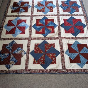 Op de afbeelding: Een handgemaakte quilt met een windmolenontwerp. De quilt heeft negen vierkanten, elk met een centraal windmolenpatroon in tinten blauw, rood en bloemenstoffen, tegen een witte achtergrond. De rand is een bloemenpatroon.