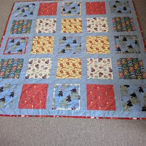 Könnte beinhalten: Ein Patchwork-Quilt mit hellblauem Hintergrund und rotem Rand. Der Quilt zeigt quadratische Blöcke mit verschiedenen Mustern, darunter Figuren in schwarzen Roben, Wappen und andere thematische Designs. Der Quilt ist wahrscheinlich handgefertigt.