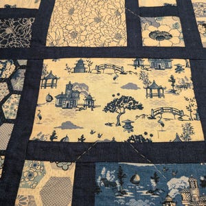 Peut inclure: Une courtepointe patchwork avec divers motifs d'inspiration japonaise. La courtepointe pr&eacute;sente des blocs rectangulaires et hexagonaux avec des images de carpes ko&iuml;, de motifs floraux et de paysages sereins avec des pagodes, des ponts et des arbres. Les blocs sont bord&eacute;s de tissu bleu marine.