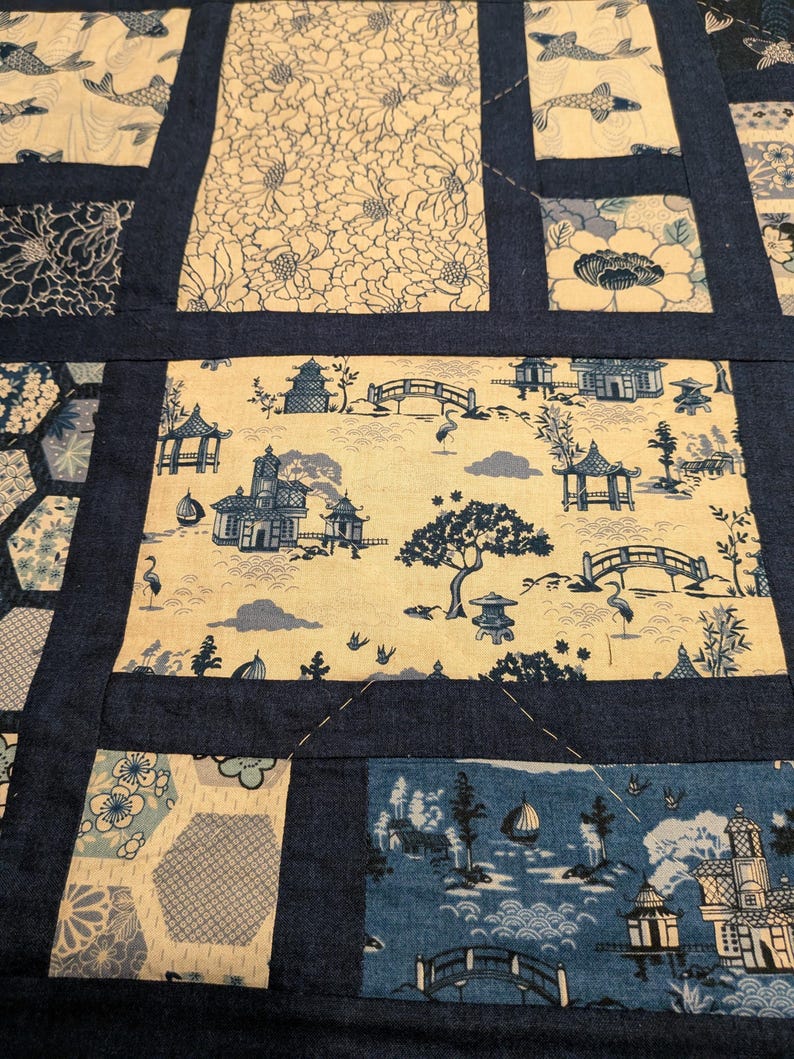 Peut inclure: Une courtepointe patchwork avec divers motifs d'inspiration japonaise. La courtepointe pr&eacute;sente des blocs rectangulaires et hexagonaux avec des images de carpes ko&iuml;, de motifs floraux et de paysages sereins avec des pagodes, des ponts et des arbres. Les blocs sont bord&eacute;s de tissu bleu marine.