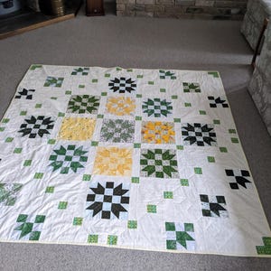 Op de afbeelding: Een handgemaakte quilt met een witte achtergrond en een herhalend patroon van vierkanten. De vierkanten hebben stervormige ontwerpen in zwart, groen en geel, met kleinere groene vierkanten verspreid. De quilt is ongeveer 178 cm bij 203 cm groot.