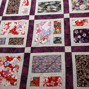 May include: A handmade quilt with a patchwork design. The quilt features a grid pattern with squares and rectangles in various floral and patterned fabrics. The dominant colours are red, purple, and white, creating a vibrant and textured appearance.