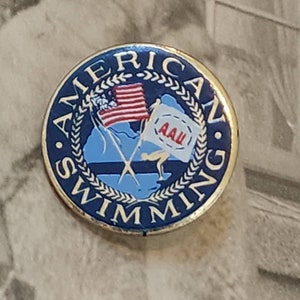 Peut inclure: Une épingle bleue et dorée avec les mots "American Swimming" autour du bord. L'épingle représente un nageur, un drapeau américain et les lettres "AAU" dans un cercle blanc.