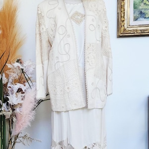 May include: A cream-colored vintage outfit on a mannequin, including a long skirt, a jacket with embroidered details, and a matching top. A straw hat sits atop the mannequin's head. Black heels are arranged at the base of the mannequin.