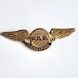 Puede incluir: Un pin dorado de la FAA de Singapur con alas extendidas. El pin presenta las letras "FAA" en el centro, rodeadas por la palabra "Singapur" en una banda azul. Las alas tienen plumas detalladas.